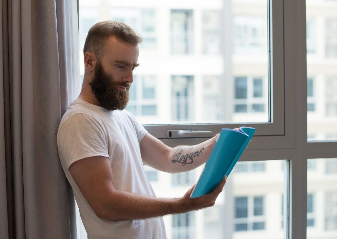 Un homme barbu lit un document bleu près d'une fenêtre lumineuse, peut-être pour des informations sur Eco Stores.