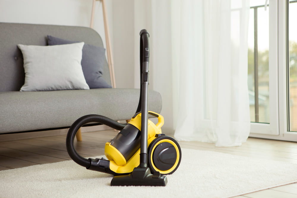 Aspirateur multifonction jaune et noir posé sur un tapis blanc dans un salon moderne.