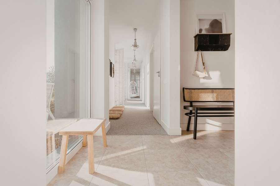 Un long couloir blanc lumineux avec des idées d'aménagement minimalistes, un banc en bois clair, une console en rotin et des suspensions vintage.