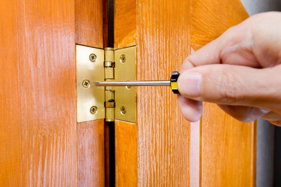 Une main visse une charnière en laiton sur une porte en bois, illustrant comment blinder les paumelles d'une porte d'entrée pour empêcher le dégondage.