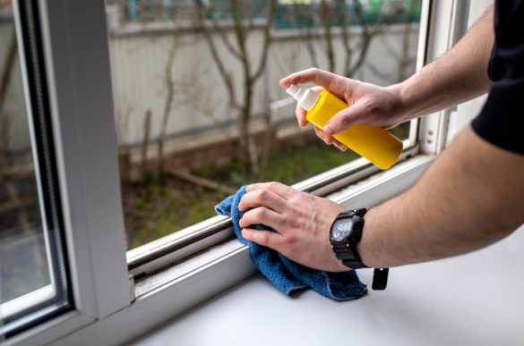 Un homme nettoie le cadre d'une fenêtre en PVC blanc avec un chiffon bleu et un spray jaune, illustrant une étape du nettoyage pour savoir comment nettoyer menuiserie pvc jaunie.